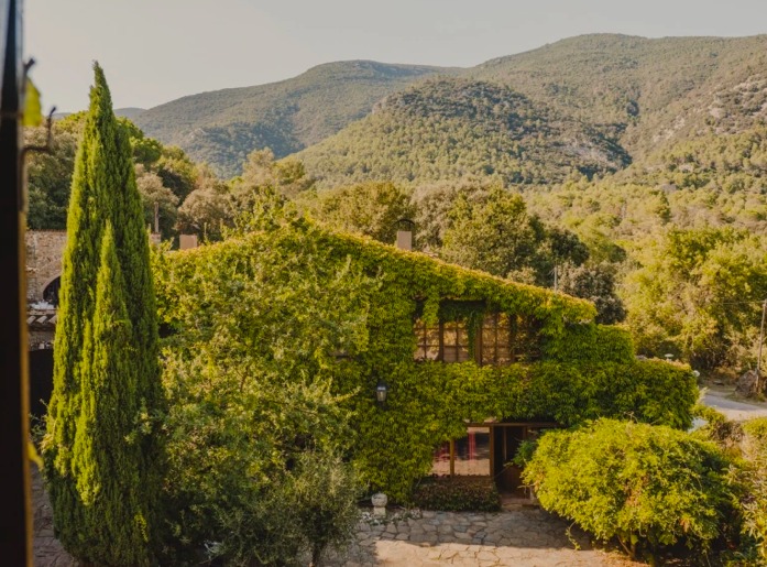A restored stone masia covered in ivy, surrounded by cypress trees and the wooded hills of La Garrotxa